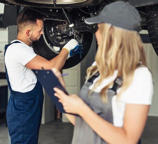 Un mécanicien effectue une inspection sous un véhicule pendant qu’une collègue note les informations, illustrant des services automobiles complémentaires.